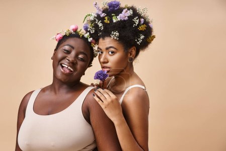 sonrientes mujeres afroamericanas con flores en el pelo posando en ropa interior en beige, además de belleza tamaño