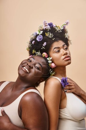 charming african american women with flowers in hair posing in underwear on beige, plus size beauty