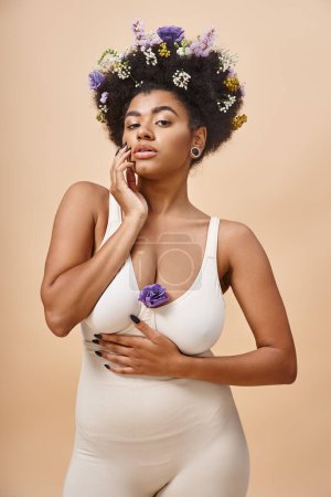 sensual mujer afroamericana en lencería posando con flores en pelo sobre beige, belleza natural