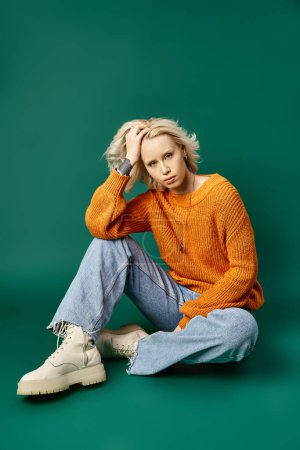 tattooed blonde woman in mustard yellow sweater and denim jeans sitting on turquoise background