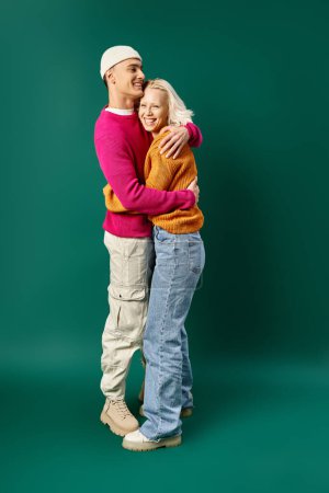 happy couple in winter attire, man in beanie hugging blonde girlfriend on turquoise, winter fashion