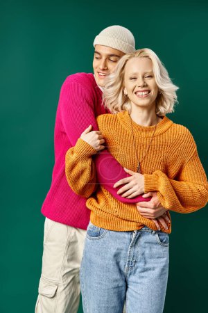 couple in winter sweaters, man in beanie hugging happy girlfriend on turquoise, winter fashion