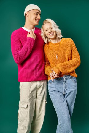 fröhliches junges Paar in Winterpullovern, lachend und vor türkisfarbenem Hintergrund in die Kamera blickend