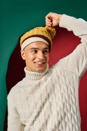 joyful man in white sweater wearing yellow mustard bobble hat on turquoise background, winter trends