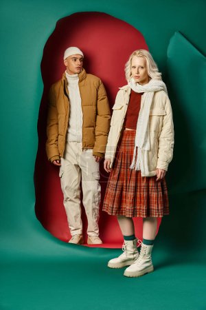 handsome man in beanie hat and puffer jacket looking at blonde woman on red with turquoise backdrop