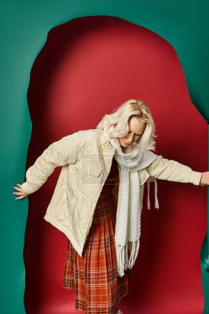young blonde woman in stylish winter attire ripping turquoise paper and posing on red backdrop