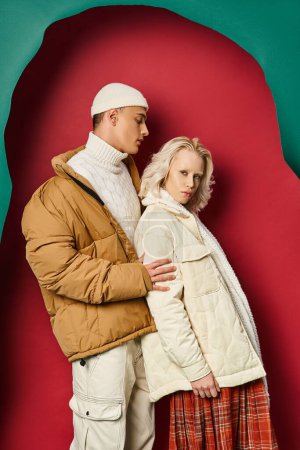 young man and woman in stylish winter jackets posing together on torn turquoise and red background