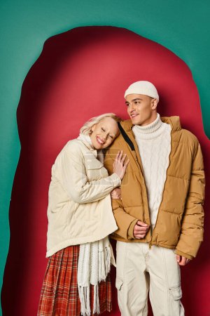 cheerful woman in winter jacket hugging stylish boyfriend near torn turquoise and red background