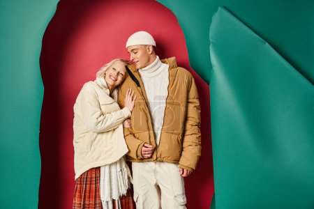 pleased woman in winter jacket hugging happy boyfriend near torn turquoise and red background