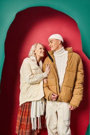 happy woman in winter jacket hugging cheerful boyfriend near torn turquoise and red background