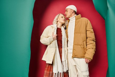 stylisches junges Paar in stylischer Winteroberbekleidung, das sich vor zerrissenem türkis-roten Hintergrund umarmt