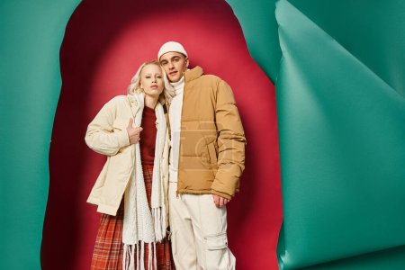 stylish young couple in warm winter outerwear posing together near torn turquoise and red backdrop