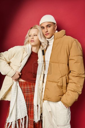 stylish man and woman in warm winter outerwear posing together on red background, winter fashion