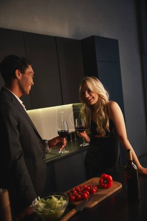cheerful multicultural couple in evening attires with wine glasses in hands smiling at each other