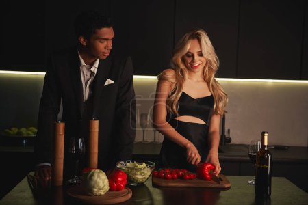 jolly attractive woman in black dress cutting bell pepper next to her african american boyfriend