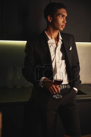 gut aussehender afrikanisch-amerikanischer Mann im eleganten Anzug posiert mit einem Glas Rotwein und schaut weg