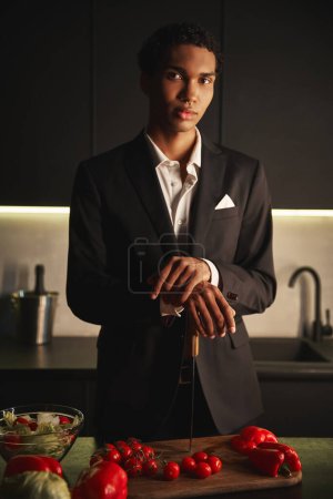 gut aussehender junger Mann im eleganten schwarzen Anzug posiert in seiner Küche und blickt direkt in die Kamera