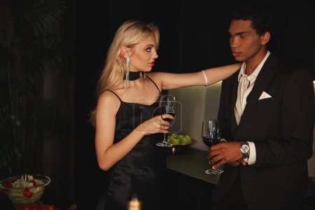 Téléchargez les photos : Séduisant couple sensuel diversifié dans des vêtements élégants tenant des verres à vin et se regardant - en image libre de droit