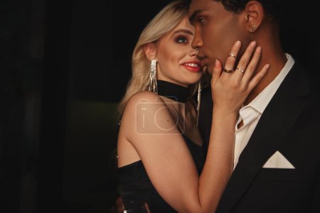 Photo for Joyous beautiful woman in black dress smiling at her african american boyfriend, hand on cheek - Royalty Free Image