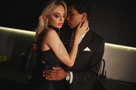 Téléchargez les photos : Séduisant couple multiculturel sensuel avec des accessoires dans des vêtements chics étreignant amoureusement dans la cuisine - en image libre de droit