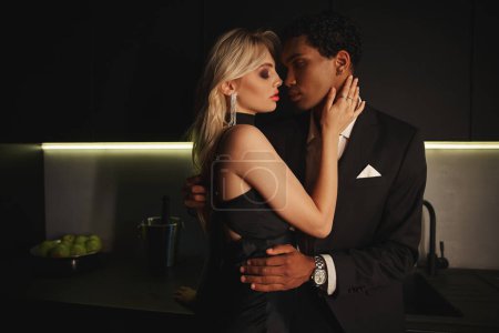 Téléchargez les photos : Séduisante sensuelle couple diversifié en soirée chic habille étreinte tandis que dans la cuisine - en image libre de droit