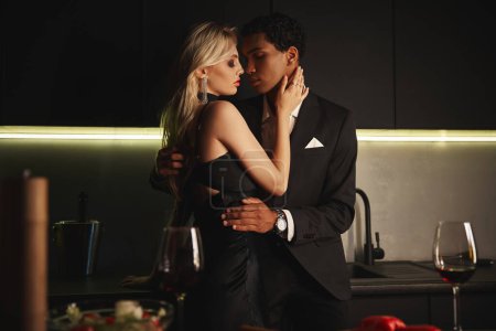 attraktive junge multiethnische Paar in eleganten stilvollen Outfits umarmen liebevoll, während in der Küche