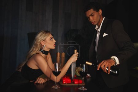 Téléchargez les photos : Belle femme avec couteau à la main en regardant son petit ami afro-américain qui verse du vin rouge - en image libre de droit