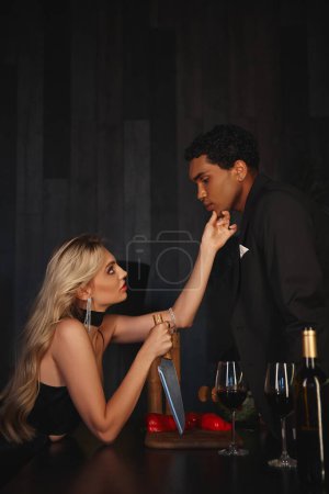 hombre afroamericano guapo en traje elegante mirando a su hermosa novia sosteniendo cuchillo