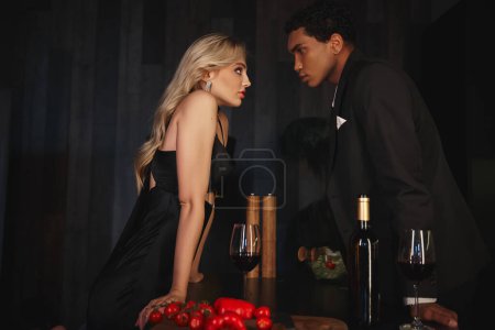 atractivo hermosa pareja multirracial en elegantes trajes mirándose entre sí mientras que en la cocina