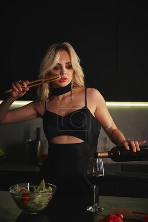 attraktive blonde junge Frau im schwarzen Kleid schenkt etwas Rotwein ein und isst Salat mit Stäbchen