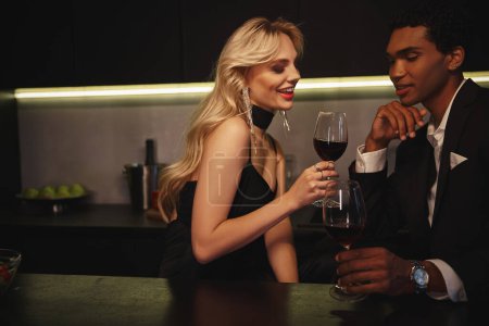 joyeux couple multiculturel aimant dans des vêtements chics boire du vin rouge et parler dans la cuisine