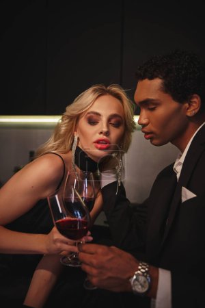 Téléchargez les photos : Jeune couple multiculturel sensuel en soirée habille cliquetis verres de vin rouge dans la cuisine - en image libre de droit