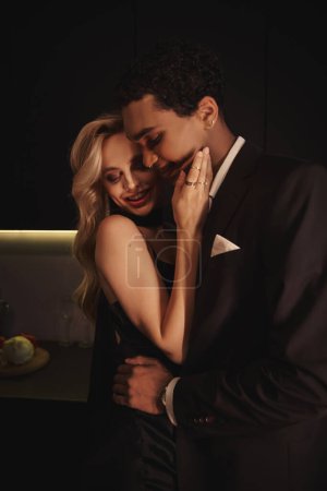 Photo for Joyful multicultural young couple in elegant evening attires hugging sensually and smiling at home - Royalty Free Image