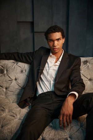 handsome relaxed african american male model in chic suit sitting on sofa and looking at camera