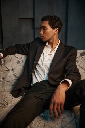 schöner junger attraktiver afrikanisch-amerikanischer Mann im schwarzen Anzug sitzt auf dem Sofa und schaut weg