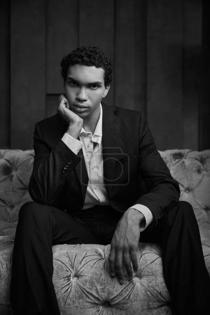 foto en blanco y negro del hombre afroamericano elegante sentado en el sofá y mirando a la cámara