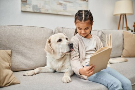 glückliches Mädchen sitzt auf Sofa und liest Buch neben Labrador im modernen Wohnzimmer, tierischer Begleiter