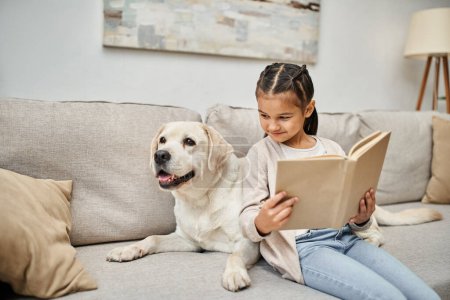 fröhliches Mädchen sitzt auf Sofa und liest Buch neben Labrador im modernen Wohnzimmer, tierischer Begleiter