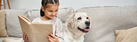 Téléchargez les photos : Fille heureuse assise sur le canapé et le livre de lecture près de labrador dans le salon moderne, bannière compagnon - en image libre de droit
