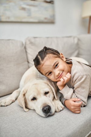portrait of happy kid smiling near cute labrador in modern living room, furry friend and child