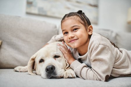 portrait of happy girl smiling near cute labrador in modern living room, domestic animal and kid