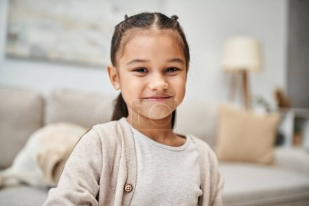 Téléchargez les photos : Portrait de heureuse fille d'âge élémentaire avec des cheveux bruns souriant à la caméra dans un appartement moderne - en image libre de droit