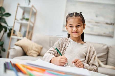 Porträt eines glücklichen Mädchens im Grundalter, das mit Farbstift zeichnet und in einer modernen Wohnung lächelt