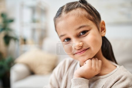 Téléchargez les photos : Portrait de fille d'âge élémentaire souriante avec des cheveux bruns regardant la caméra dans un appartement moderne - en image libre de droit