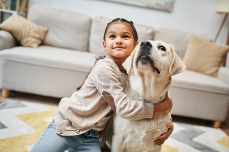 happy cute elementary age girl looking up and hugging labrador in modern apartment, pet and child