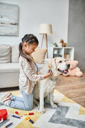 glückliches Mädchen in lässiger Kleidung spielt Arzt mit Labrador im modernen Wohnzimmer, Spielzeug-Verbandskasten
