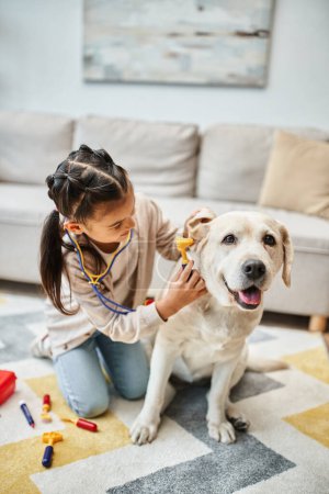Nettes Mädchen spielt Arzt mit Labrador im modernen Wohnzimmer, Spielzeug-Verbandskasten und Stethoskop