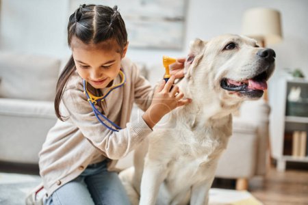 glückliches Kind in Freizeitkleidung spielt Arzt mit Labrador im modernen Wohnzimmer, Spielzeug-Stethoskop