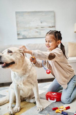 glückliches Kind in Freizeitkleidung, spielender Arzt mit Labrador im modernen Wohnzimmer, Spielzeug-Stethoskop