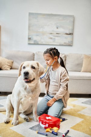 fröhliches Mädchen in lässiger Kleidung spielt Arzt mit Labrador-Hund im Wohnzimmer, Spielzeug-Verbandskasten
